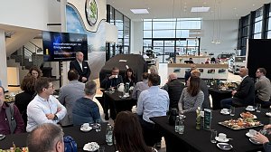 Torsten Wiesner hält einen Vortrag mit der Präsentation Herausforderungen der Energiewende. Teilnehmende sitzen an runden Tischen mit Kaffee und Frühstücksplatten.