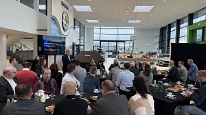 Ein Referent hält einen Vortrag mit der Präsentation Herausforderungen der Energiewende. Teilnehmende sitzen an runden Tischen mit Kaffee und Frühstücksplatten.
