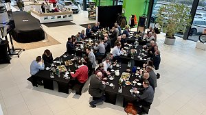 Blick von oben auf den Ausstellungsraum. Mehrere lange Tische sind mit Frühstück gedeckt. Viele Unternehmerinnen und Unternehmer sitzen und diskutieren. Links im Bild ein abgedecktes Fahrzeug.
