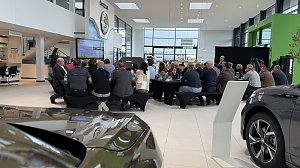 Rückansicht der Gäste, die an Frühstückstischen sitzen und einer Präsentation zuhören. Links im Bild die Rednerin, rechts der große Škoda-Schriftzug an der Wand.