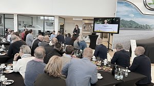 Mehrere Dutzend Gäste sitzen an langen Tischen und schauen auf die Rednerin. Auf dem Bildschirm ist die Abschlussfolie Fragen? zu sehen. Im Vordergrund Teller und Getränke.