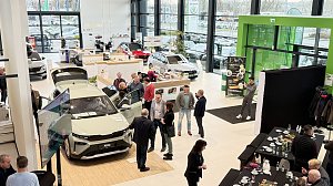 Im hellen Ausstellungsraum des Škoda Autohauses steht ein neuer Škoda Elroq mit geöffneten Türen und Kofferraum. Mehrere Gäste betrachten das Fahrzeug, einige unterhalten sich in kleinen Gruppen. Im Hintergrund sind weitere Autos sowie der Empfangsbereich mit der Beschilderung Information und Kasse zu sehen. Rechts am Rand ein Buffetbereich mit leeren Tassen und Tellern, daneben ein schwarzer Fotohintergrund.