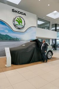 Ein Škoda Elroq ist noch mit einem schwarzen Tuch abgedeckt. Ein Mitarbeiter zieht die Abdeckung teilweise zur Seite. Das große Škoda-Logo hängt an der Wand.