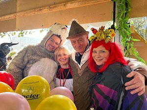 "Vier Personen in farbenfrohen Kostümen – darunter ein Bärenkostüm, ein Dirndl und ein Fantasiekostüm mit roter Perücke – stehen beieinander und lächeln in die Kamera, umgeben von bunten Luftballons.