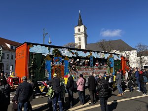 Karnevalswagen mit Burg- und Wolkenmotiven fährt durch eine Innenstadt, Menschen stehen am Straßenrand und schauen zu, im Hintergrund eine Kirche mit Turm.