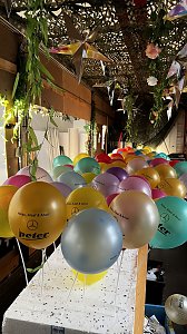 "Reihe bunter Luftballons mit der Aufschrift ‚Helau, Alaaf & Ahoi!‘ und dem Autohaus-Peter-Logo, befestigt an weißen Stäben, unter einer mit Pflanzen und Sternen dekorierten Decke.