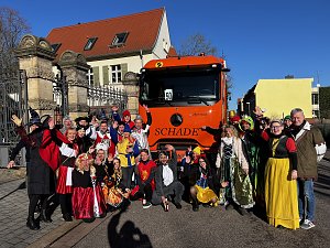 "Gruppe von kostümierten Menschen in Märchen- und Tierverkleidungen posiert vor einem orangefarbenen LKW mit der Aufschrift ‚SCHADE‘.