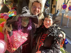 rei verkleidete Personen – eine Hexe mit grünem Gesicht und spitzem Hut, eine Person im weißen Bärenkostüm und eine als Maleficent verkleidete Frau – posieren lachend, bunte Luftballons im Hintergrund.