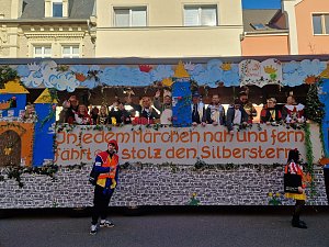Seitenansicht des bunt gestalteten Karnevalswagens mit dem Schriftzug ‚In jedem Märchen nah und fern fährt er stolz den Silberstern‘, mehrere verkleidete Menschen stehen darauf, zwei Personen in Narrenkostümen davor.