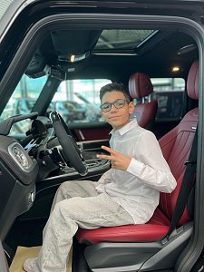 Jamil aus ‚The Voice Kids‘ sitzt auf dem Fahrersitz eines schwarzen Mercedes mit roter Lederausstattung, lächelt in die Kamera und zeigt ein Peace-Zeichen.