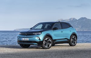Türkis-schwarzer Opel Mokka Electric steht am Meer, Blick schräg von vorn links, Berge im Hintergrund.