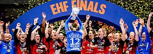 Das Bild zeigt ein Handballteam, das gerade den Sieg bei den EHF Finals feiert. In der Mitte hebt die Torhüterin mit der Rückennummer 12 („Eckerle“) den Pokal über den Kopf, während gelbe Konfettistreifen durch die Luft wirbeln. Ihre Mitspielerinnen und das Trainerteam jubeln, klatschen und reißen die Arme in die Höhe. Alle tragen Medaillen und schwarze-rote Trikots, einige blaue Torwarttrikots. Im Hintergrund ist ein großer blauer Torbogen mit der Aufschrift „EHF FINALS“ zu sehen.