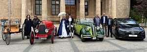 Gruppenfoto mit Fahrzeugen verschiedener Epochen: Vor einem historischen Gebäude stehen vier Autos  von einer Kutsche mit Holzaufbau über einen roten Oldtimer, einen grünen Oldtimer-Cabrio bis zu einem modernen schwarzen Mercedes. Davor posieren Personen in historischer Kleidung und moderner Garderobe.