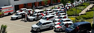 Gruppenfoto mit Fahrzeugflotte: Mehrere neue und bestehende Fahrzeuge des DRK Nordhausen, überwiegend weiß mit rotem Kreuz, stehen in einem Innenhof. DRK-Mitarbeitende in roten Shirts posieren neben den Autos, im Vordergrund ein dunkelblauer Transporter.