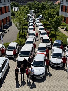 Fuhrparkaufstellung von oben: Reihe von DRK-Fahrzeugen, flankiert von Mitarbeitenden in roten Shirts. Im Vordergrund werden symbolisch Fahrzeugschlüssel und Blumen übergeben.