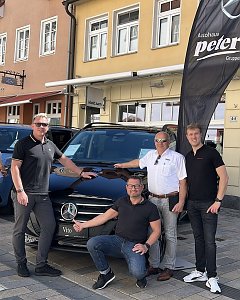 Vier Männer posieren vor einem schwarzen Mercedes Vito auf einer Kopfsteinpflasterstraße in Heiligenstadt. Links und rechts im Hintergrund stehen weitere Fahrzeuge, im Hintergrund sind mehrstöckige Altstadthäuser mit Schaufenstern zu sehen. Neben dem Auto weht eine schwarze Autohaus-Peter-Flagge.