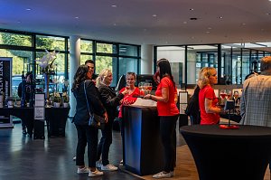 Mehrere Frauen, teils in roten She’s Mercedes-T-Shirts, teils in schwarzen Outfits, stehen an einem Stehtisch mit Gläsern und unterhalten sich in einem hellen Autohaus.
