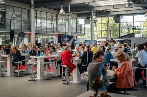 Publikum im Showroom: Viele Frauen sitzen an hohen weißen Tischen in einem großen, hellen Autohaus mit Glasfront, trinken und unterhalten sich. Im Hintergrund sind Mercedes-Werbematerialien und Fahrzeuge sichtbar.