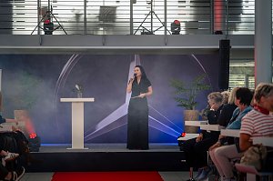 Christina Dost spricht auf einer Bühne mit Mercedes-Logo im Hintergrund, rotem Teppich davor und Publikum auf beiden Seiten.