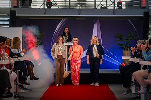 Drei Frauen laufen in modischen Outfits über einen roten Teppich auf einer Bühne mit Mercedes-Logo, während eine Moderatorin am Pult steht. Das Publikum applaudiert.