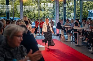 Frau mit kurzem blondem Haar, weißer Jacke und langem, dunkelblauem Kleid mit weißen Punkten läuft über den roten Teppich.