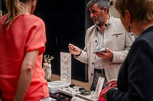 Ein Mann in heller Jacke präsentiert Schmuckstücke an einem Stand, während zwei Frauen interessiert zuhören. Auf dem Tisch liegen Ketten, Armbänder und Ringe.