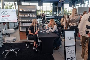 Zwei Frauen sitzen an einem Tisch und unterhalten sich; im Hintergrund steht ein Regal mit Kosmetikprodukten und ein Bildschirm mit der Aufschrift She’s Mercedes Ladies Night 22. Mai 2025. Daneben stöbern Besucher an einem Kleiderständer.