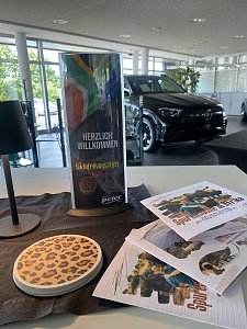 Dekoration zum Südafrikanischen Abend im Autohaus Peter: Ein Aufsteller mit der Aufschrift ‚Herzlich Willkommen zum Südafrikanischen Abend‘ und südafrikanischer Flagge, daneben Flyer mit Safari-Motiven und ein Untersetzer mit Leopardenmuster. Im Hintergrund steht ein schwarzer Mercedes im Showroom.