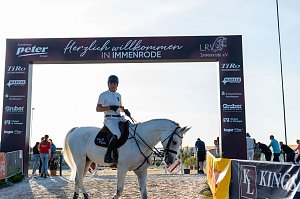 Ein Reiter in weißem T-Shirt und schwarzem Helm sitzt auf einem weißen Pferd unter einem Begrüßungsbogensorenn mit der Aufschrift ‚Herzlich willkommen in Immenrode‘. Der Bogen ist mit Logos verschiedener Spo versehen. Im Hintergrund sind mehrere Personen und ein sonniger Himmel zu sehen.