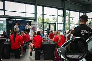 Helmut Peter, Trainer Herbert Müller und Robert Böhm (von links nach rechts) stehen auf einer Bühne beim Medien-Tag des Thüringer HC in Erfurt. Im Vordergrund sitzen Spielerinnen in roten Trikots an Tischen und hören zu. Hinter den Männern steht ein THC-Banner und durch die großen Glasfenster sind Autos und Bäume zu sehen.