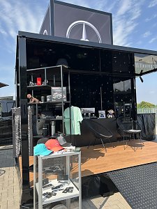 Schwarzer Mercedes-Stand mit großem Mercedes-Logo auf dem Dach. Im Vordergrund ein Glasdisplay mit Schlüsselanhängern und Caps in Schwarz, Rot und Türkis mit Mercedes-Stern. Dahinter Regale mit Merchandise-Artikeln wie Thermobecher, Modellautos, Kleidung und einem Stofftier. Zwei schwarze Stühle und ein Tisch stehen auf der hölzernen Ausstellungsfläche.