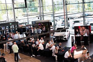 Eine große Halle mit vielen Gästen, die an schwarzen Tischen sitzen oder an Stehtischen stehen. Ein weißer Mercedes ist ausgestellt, dahinter eine Bar und ein Bildschirm mit dem Mercedes-Logo.