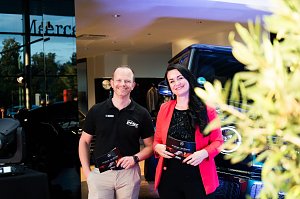 Eine Frau im roten Blazer (Christina Dost) und ein Mann im schwarzen Polohemd (Carsten Dost) stehen nebeneinander und lächeln. Beide halten Moderationskarten, im Hintergrund ein Mercedes-Geländewagen.