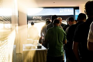 Eine Gruppe Männer steht in einer Schlange am Buffet. Sonnenlicht fällt durch die Tür, im Hintergrund ein Pavillon mit Autohaus-Peter-Logo.