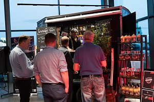Mehrere Männer stehen an einem Gin-Stand mit schwarzem Verkaufswagen. Auf Schildern steht Luzifer Gin und Gin-Cocktails. Regale mit Flaschen sind daneben aufgestellt.