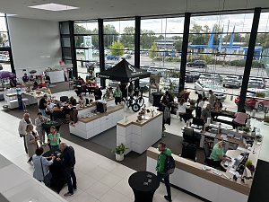 Blick von oben in den hellen Ausstellungsraum des Autohauses Autowelt Peter während der Feier zu 130 Jahren Škoda. Besucherinnen und Besucher sitzen an Tischen, basteln und stricken, andere informieren sich an Ständen, darunter ein SCOTT-Zelt mit Fahrrädern.