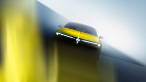 Ein gelbes Auto ist frontal und leicht von unten fotografiert. Das Fahrzeug steht in einer nebeligen, lichtdurchfluteten Umgebung. Die dominante Frontleiste mit schmalen LED-Scheinwerfern bildet eine horizontale Lichtlinie. In der Mitte befindet sich ein kreisförmiges Markenemblem. Die Aufnahme wirkt dynamisch und futuristisch durch starke Unschärfen und Lichtreflexe.