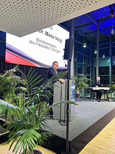 Redner am Pult auf der Bühne der Automobil Messe Erfurt, Leinwand: Dr. Mario Voigt, Ministerpräsident Thüringen, rechts ein Keyboardspieler.