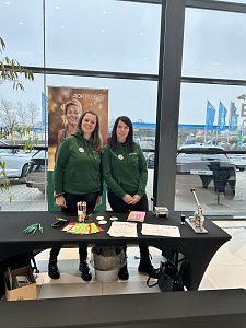 Zwei Frauen in grünen Hoodies stehen an einem Infotisch mit Stiften und Flyern, dahinter ein AOK-Plus-Banner.