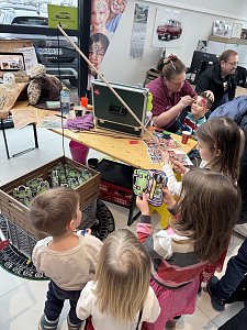 Mehrere Kinder stehen an einem Aktionsstand, während eine Frau einem Jungen das Gesicht schminkt. Auf dem Tisch liegen Farben, Flyer und Zubehör.