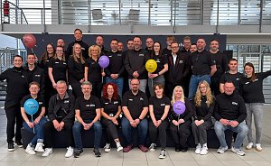 Man sieht ein Teamfoto mit vielen Menschen auf und vor einer kleinen B&uuml;hne. Alle haben das gleiche T-Shirt an, manche halten bunte Luftballons hoch.