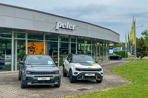 Au&szlig;enansicht des Autohauses Peter in Halle Neustadt/ G&ouml;ttinger Bogen. Im Vordergrund steht ein grauer und ein wei&szlig;er Jeep. Rechts stehen Fahnenmasten auf einer Gr&uuml;nfl&auml;che. Im Hintergrund ist das Autohaus mit einer gro&szlig;en Glasfront.