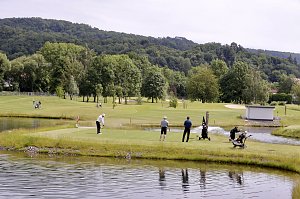 Golfer spielen an einem Abschlag mit Wasserhindernis vor bewaldeter Kulisse.