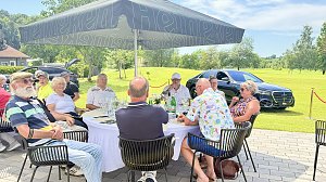 Gäste sitzen an einem Tisch unter Sonnenschirmen mit Blick auf den Golfplatz und ausgestellte Fahrzeuge.