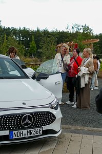 Eine Gruppe von Frauen, die sich im Freien unter bewölktem Himmel versammelt haben und aufmerksam einen weißen Mercedes-Benz CLA betrachten.