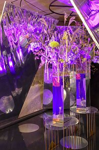 Hohe Glasvasen mit floralen Arrangements, die auf kleinen runden Tischen unter intensivem, lilafarbenem Event-Licht vor spiegelnden Wänden stehen.