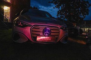 Die Frontansicht eines Mercedes-Benz, der in der Dämmerung im Freien steht und dessen Kühlergrill sowie das Markenlogo lila beleuchtet sind.