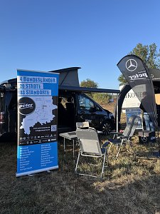 Stand vom Autohaus Peter. Im Vordergrund ein gro&szlig;es blaues Werbebanner mit Firmeninfos. Dahinter eine V-Klasse Marco Polo mit ge&ouml;ffnetem Campingdach. Davor stehen ein Tisch und zwei Campingst&uuml;hle. Rechts eine Mercedes-Fahne und ein Pavillon.