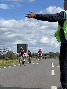 Radstrecke beim Triathlon. Im Vordergrund weist ein Streckenposten mit ausgestrecktem Arm den Weg. Dahinter fahren mehrere Radfahrer mit Helmen auf einer asphaltierten Stra&szlig;e. Im Hintergrund ist ein bew&ouml;lkter Himmel zu sehen.