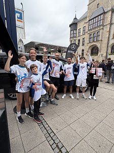 Man sieht unser Firmenlauf-Team nach dem Lauf, vor einem schwarzen EQT. Dahinter steht eine schwarze Beachflag mit unserem Logo in Weiß und dem weißen Mercedes-Benz Stern. Im Hintergrund sieht man das Dessauer Rathaus.